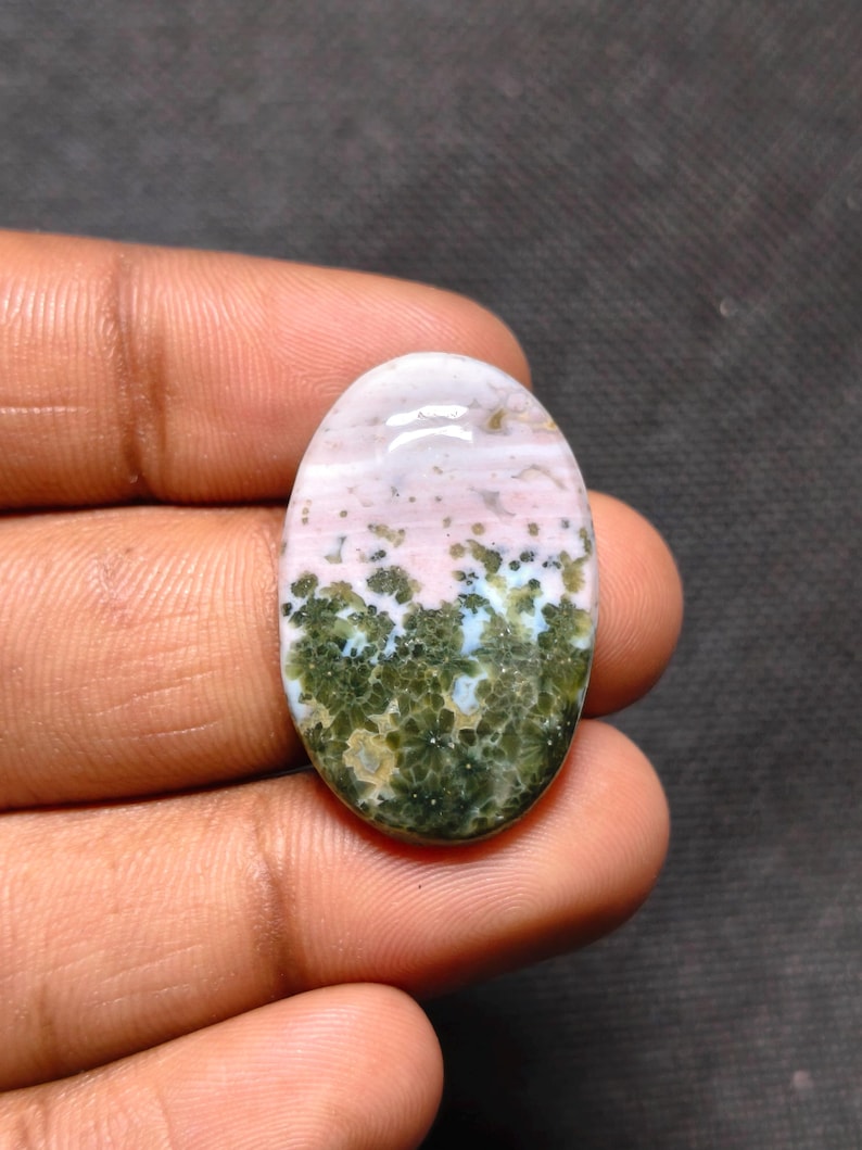May include: An oval-shaped cabochon gemstone featuring a smooth, polished surface. The stone displays a distinct color gradient, transitioning from a soft, pale pink at the top to a deep, mossy green at the bottom with intricate floral-like patterns.