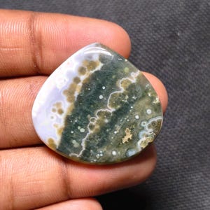 May include: A polished, triangular-shaped ocean jasper gemstone featuring a smooth surface with a color palette of deep forest green, olive, and creamy white, displaying distinct circular orbicular patterns and translucent sections.