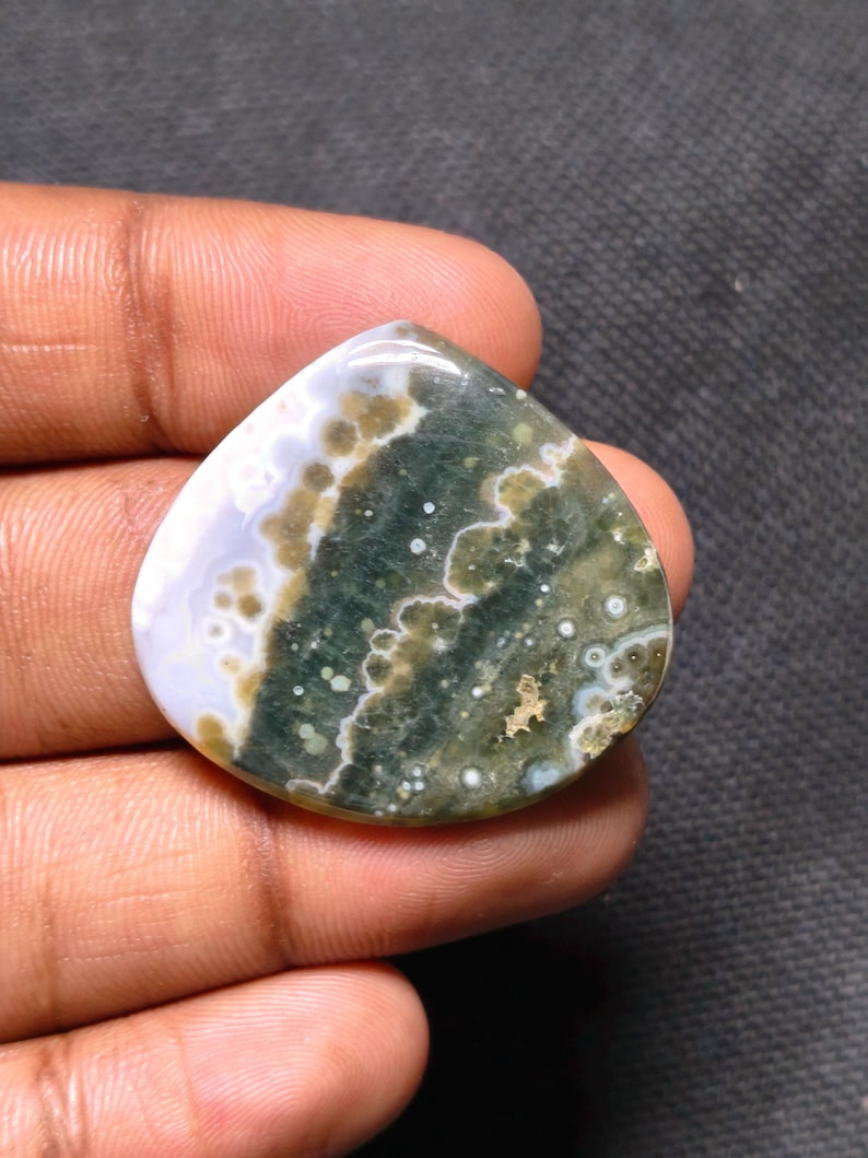 May include: A polished, triangular-shaped ocean jasper gemstone featuring a smooth surface with a color palette of deep forest green, olive, and creamy white, displaying distinct circular orbicular patterns and translucent sections.