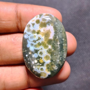 May include: An oval-shaped ocean jasper gemstone featuring a polished surface with a complex pattern of mossy green, cream, and soft blue circular orbs and inclusions set against a dark, deep forest green background.