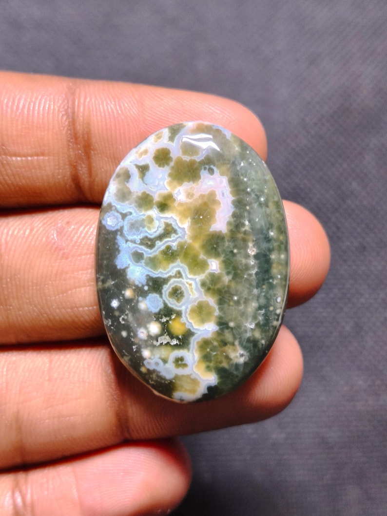 May include: An oval-shaped ocean jasper gemstone featuring a polished surface with a complex pattern of mossy green, cream, and soft blue circular orbs and inclusions set against a dark, deep forest green background.