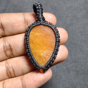 Op de afbeelding: Een druppelvormige hanger met een heldere oranje edelsteen, vakkundig gevat in een zwart macramé-ontwerp. De hanger hangt aan een geweven lus en toont een ambachtelijke esthetiek. De kleur van de edelsteen varieert van licht- tot donkeroranje.
