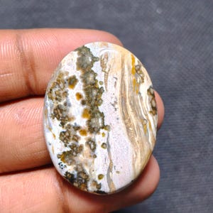 May include: An oval-shaped polished gemstone featuring a complex pattern of white, beige, and brown swirls. The surface displays dark mossy inclusions and golden-yellow spots distributed across the smooth, rounded stone face.
