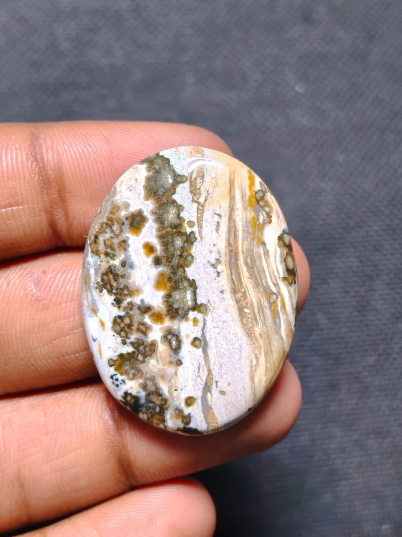 May include: An oval-shaped polished gemstone featuring a complex pattern of white, beige, and brown swirls. The surface displays dark mossy inclusions and golden-yellow spots distributed across the smooth, rounded stone face.