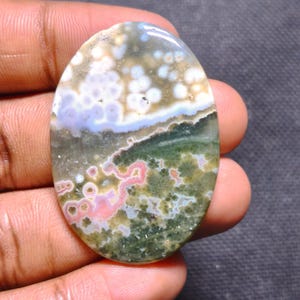 May include: An oval-shaped ocean jasper gemstone featuring a smooth, polished surface with a complex pattern of white, light blue, pink, and deep green orbicular inclusions and moss-like formations throughout the stone.