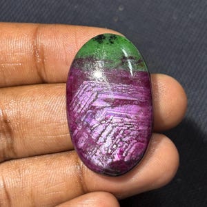May include: An oval-shaped cabochon gemstone featuring a vibrant magenta and deep purple lower section with a distinct crystalline texture, contrasted by a smooth, solid green upper section with small dark inclusions throughout the surface.