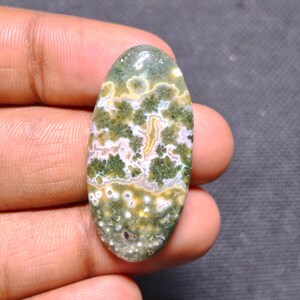 May include: An oval-shaped polished gemstone featuring a complex pattern of moss green, cream, and white inclusions. The stone displays distinct floral-like clusters and small circular spots throughout its smooth, glossy surface.
