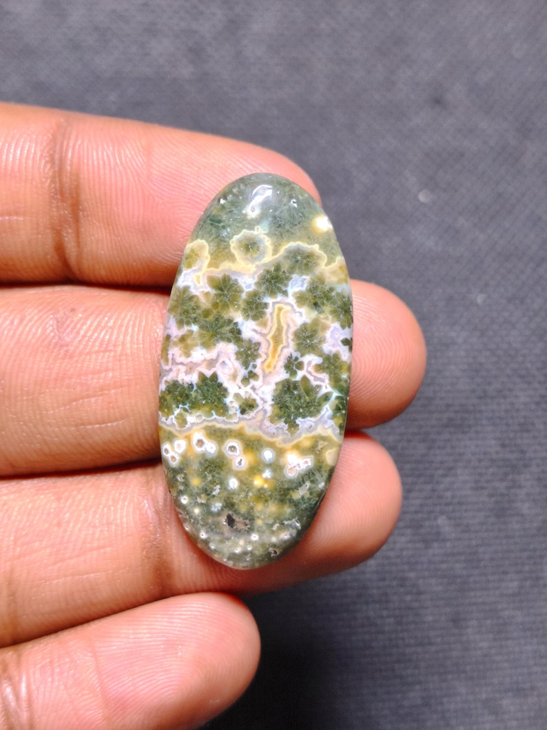 May include: An oval-shaped polished gemstone featuring a complex pattern of moss green, cream, and white inclusions. The stone displays distinct floral-like clusters and small circular spots throughout its smooth, glossy surface.