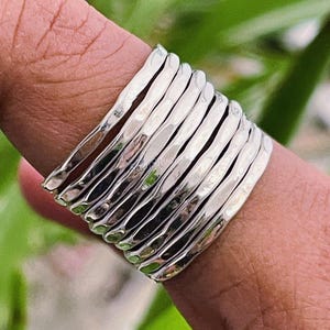 May include: A silver ring with a hammered texture, made up of several bands. The ring is on a finger, with a blurred green background.