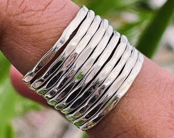 May include: A silver ring with a hammered texture, composed of multiple bands. The ring is worn on a finger, with a green and blurred background.