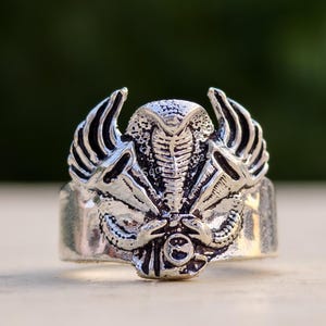 Può includere: Un anello in metallo color argento con un rilievo dettagliato di un cobra con il cappuccio spiegato, affiancato da strutture stilizzate a forma di ala su entrambi i lati. Il design presenta texture complesse e un piccolo elemento circolare.