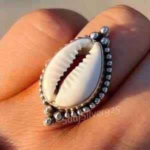 Puede incluir: Un anillo de plata con un centro de concha de cauri blanca. La concha es ovalada y está engastada en una banda de plata detallada con pequeños detalles de cuentas. El anillo está en un dedo.