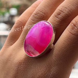 May include: A striking silver ring featuring a large, oval pink agate gemstone. The stone showcases a blend of pink shades and white marbling. This bold ring is a statement accessory. The ring's design is eye-catching and suitable for various occasions.