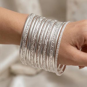 Pode incluir: Um conjunto de dez pulseiras prateadas com várias texturas e padrões, incluindo gravações florais, linhas onduladas e um design trançado. As pulseiras estão empilhadas, exibindo um acabamento metálico polido e detalhes.