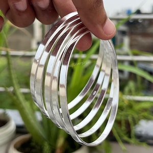 Peut inclure: Un ensemble de six bracelets argentés brillants. Les bracelets sont lisses et ronds, reflétant la lumière. Les bracelets sont empilés, créant un effet de superposition. Le fond comprend des plantes vertes.