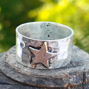 Op de afbeelding: Een zilveren ring met een gehamerde textuur en een stervormig accent. De ster heeft een zilveren centrum en goudkleurige randen. De ring is breed en ligt op een ruw, grijs oppervlak tegen een vage groene achtergrond.