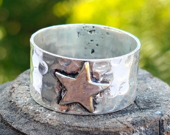 Puede incluir: Un anillo plateado con textura martillada y un adorno en forma de estrella. La estrella tiene un centro plateado y bordes dorados. El anillo es ancho y se asienta sobre una superficie gris rugosa sobre un fondo verde borroso.