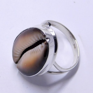 Op de afbeelding: Een zilveren ring met een natuurlijke kaurischelp. De schelp heeft bruine en crèmekleurige tinten, gezet in een gepolijste zilveren band. Het ontwerp van de ring benadrukt de unieke textuur en vorm van de schelp.