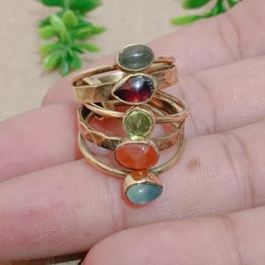 Puede incluir: Una pila de cinco anillos dorados, cada uno con una gema diferente. Las gemas son de forma ovalada o de lágrima e incluyen tonos verdes, rojos, naranjas y azules. Los anillos están apilados uno encima del otro.