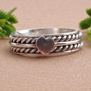 May include: A silver-colored ring with a heart-shaped center. The ring has a braided rope design around the band. Green leafy accents are on either side of the ring, set against a tan background.