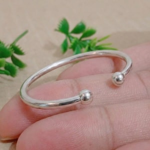 May include: A silver-colored open cuff bracelet with a smooth, rounded band. Each end of the bracelet features a small, spherical accent. The bracelet is shown against a neutral background, highlighting its simple design.