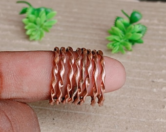 Può includere: Una collezione di anelli color rame, dal design ondulato. Gli anelli sono impilati su un dito, mostrando il loro intricato motivo strutturato. Due piccole piante decorative verdi sono sullo sfondo.