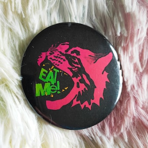 May include: A circular black pin-back button featuring a stylised hot pink graphic of a snarling animal head with its mouth open. Inside the open mouth, the text EAT ME! is printed in bright green capital letters with a jagged, distressed font.