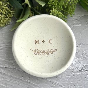May include: A white ceramic ring dish with a speckled finish. The dish features the initials "M + C" and a small leafy branch design.