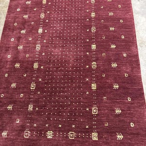 May include: A burgundy-colored rectangular rug with a repeating pattern of small squares, rectangles, and arrow-like shapes in a lighter shade. The rug has a textured surface and a symmetrical design, suitable for home decor.