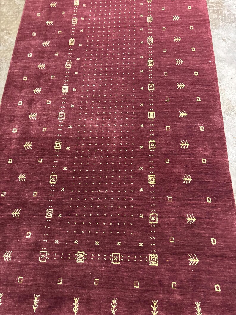 May include: A burgundy-colored rectangular rug with a repeating pattern of small squares, rectangles, and arrow-like shapes in a lighter shade. The rug has a textured surface and a symmetrical design, suitable for home decor.