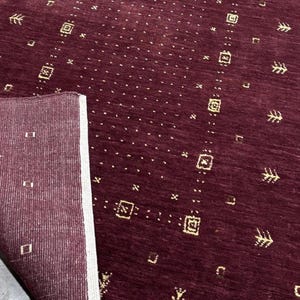 May include: A burgundy-colored rug with a geometric pattern. The rug has a rectangular shape with a folded corner, revealing a lighter-colored underside. The design includes small squares, rectangles, and arrow-like shapes in a gold color.