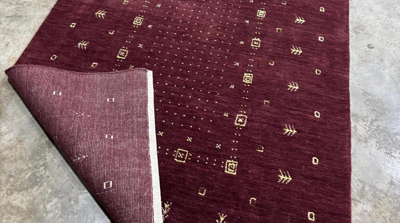 May include: A burgundy-colored rug with a geometric pattern. The rug has a rectangular shape with a folded corner, revealing a lighter-colored underside. The design includes small squares, rectangles, and arrow-like shapes in a gold color.