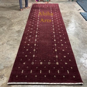 May include: A rectangular, burgundy-colored rug with a central design of small squares and dots, and a border of repeating geometric shapes. The rug has a white border and the text "Abiba Arts" in gold lettering.