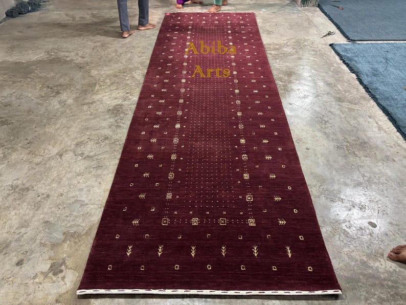 May include: A rectangular, burgundy-colored rug with a central design of small squares and dots, and a border of repeating geometric shapes. The rug has a white border and the text "Abiba Arts" in gold lettering.