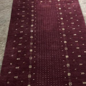 May include: A rectangular burgundy rug with a central dotted pattern and repeating geometric and stylized leaf motifs. The rug has a border with similar designs. The rug is made of a woven material.