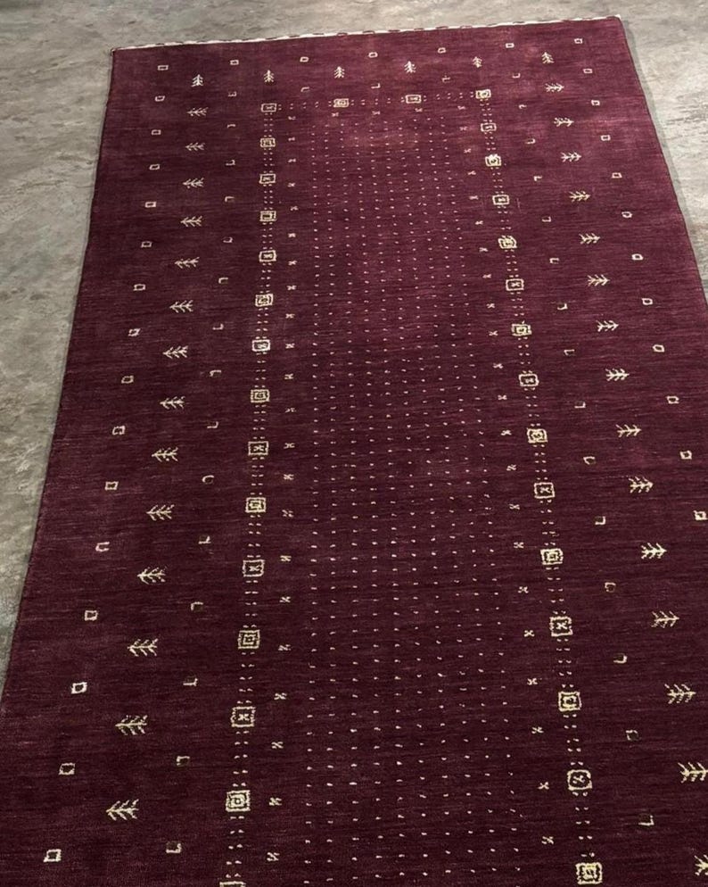 May include: A rectangular burgundy rug with a central dotted pattern and repeating geometric and stylized leaf motifs. The rug has a border with similar designs. The rug is made of a woven material.