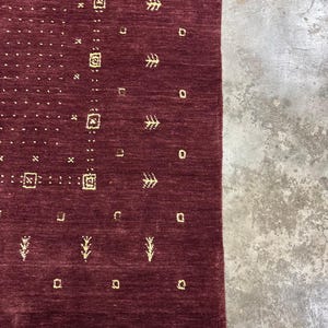 May include: A section of a burgundy-colored rug with a geometric pattern. The rug features small, repeating shapes in a lighter, golden hue, including squares, dots, and stylized tree-like designs. The edge of the rug has a white border.