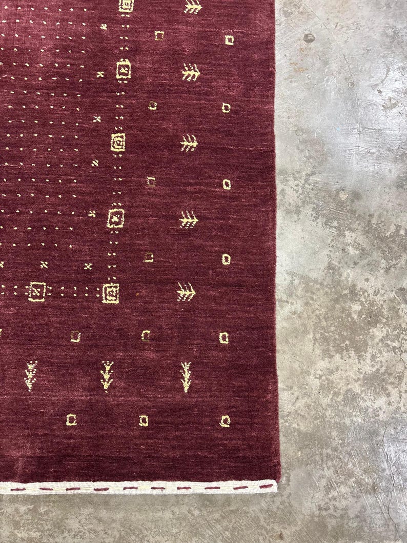 May include: A section of a burgundy-colored rug with a geometric pattern. The rug features small, repeating shapes in a lighter, golden hue, including squares, dots, and stylized tree-like designs. The edge of the rug has a white border.