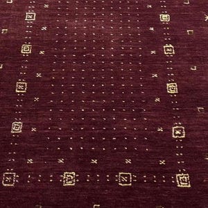 May include: A rectangular burgundy rug with a repeating pattern of small squares, arrows, and dots in a lighter shade. The rug is made of a woven material and is displayed on a concrete floor. The design is symmetrical and geometric.