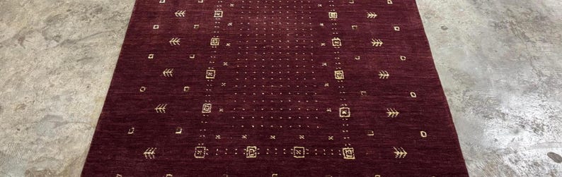 May include: A rectangular burgundy rug with a repeating pattern of small squares, arrows, and dots in a lighter shade. The rug is made of a woven material and is displayed on a concrete floor. The design is symmetrical and geometric.