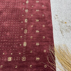 May include: A close-up of a burgundy rug with a repeating pattern of small gold squares, tree-like shapes, and dotted lines. The rug has a textured surface and a rectangular shape. The rug is partially visible, with a portion of a light-colored surface in the background.