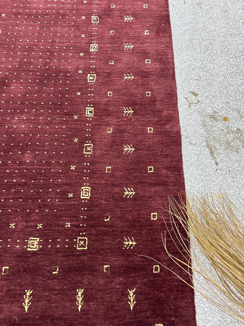 May include: A close-up of a burgundy rug with a repeating pattern of small gold squares, tree-like shapes, and dotted lines. The rug has a textured surface and a rectangular shape. The rug is partially visible, with a portion of a light-colored surface in the background.