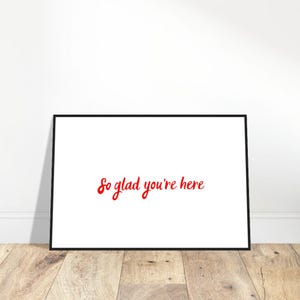 May include: A framed print with a white background and the red text "So glad you're here." The black frame contrasts with the white background. The print is displayed on a wooden floor.