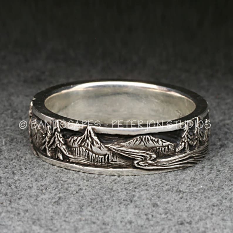 May include: A silver ring featuring a detailed relief landscape of mountains, pine trees, and a winding river. The text BANDSCAPES - PETER JON STUDIOS is engraved along the upper edge of the band. The metal has a polished finish with darkened recessed areas to highlight the intricate scenery.