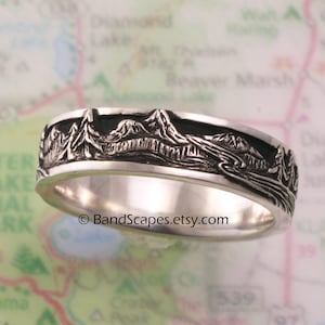 May include: A silver ring featuring a detailed relief carving of a mountain range, pine trees, and a winding river. The recessed areas of the design are darkened to provide contrast. The text BandScapes.etsy.com is printed across the center.