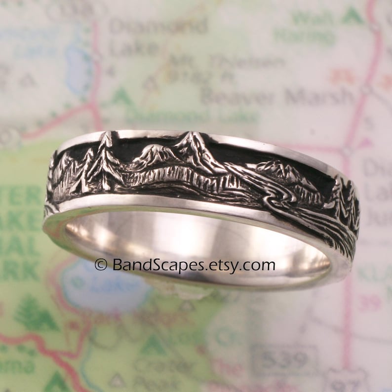 May include: A silver ring featuring a detailed relief carving of a mountain range, pine trees, and a winding river. The recessed areas of the design are darkened to provide contrast. The text BandScapes.etsy.com is printed across the center.