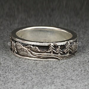 May include: A silver ring featuring a relief landscape design of mountains and pine trees with a dark oxidized finish in the recessed areas. The text BANDSCAPES - PETER JON STUDIOS is engraved along the top edge of the band.