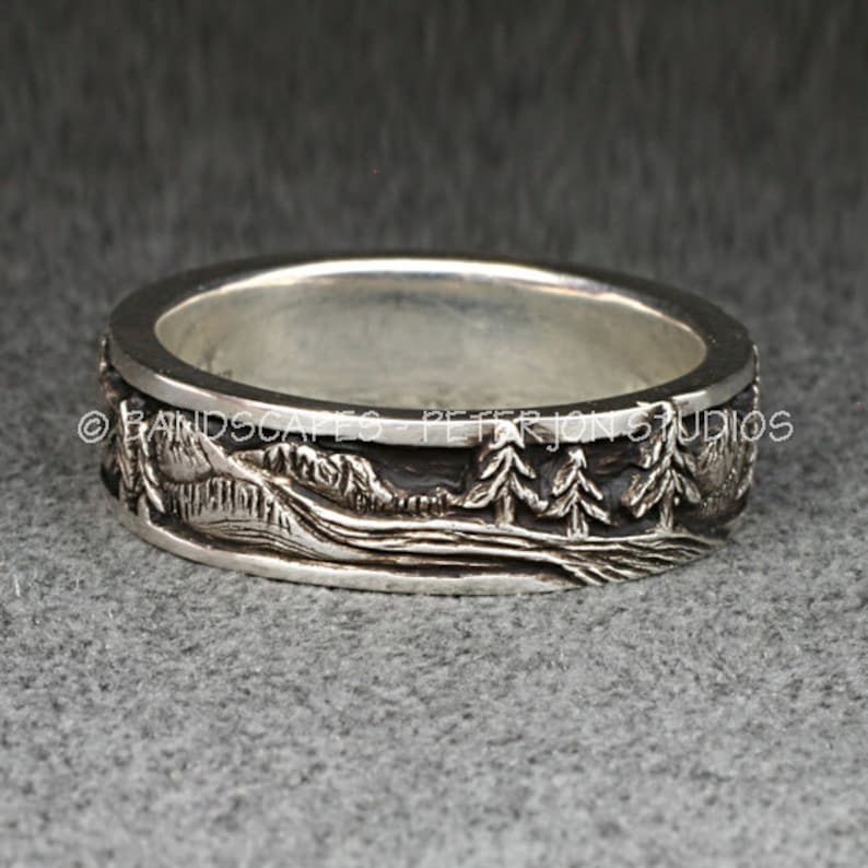 May include: A silver ring featuring a relief landscape design of mountains and pine trees with a dark oxidized finish in the recessed areas. The text BANDSCAPES - PETER JON STUDIOS is engraved along the top edge of the band.