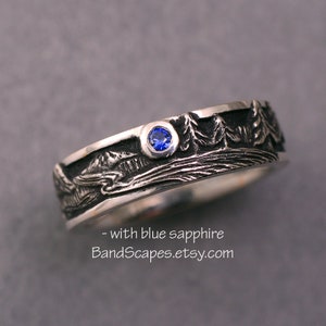 May include: A silver band ring featuring a relief landscape of trees and mountains with a single round blue sapphire set in the metal. The text - with blue sapphire BandScapes.etsy.com is printed below the ring on the gray surface.