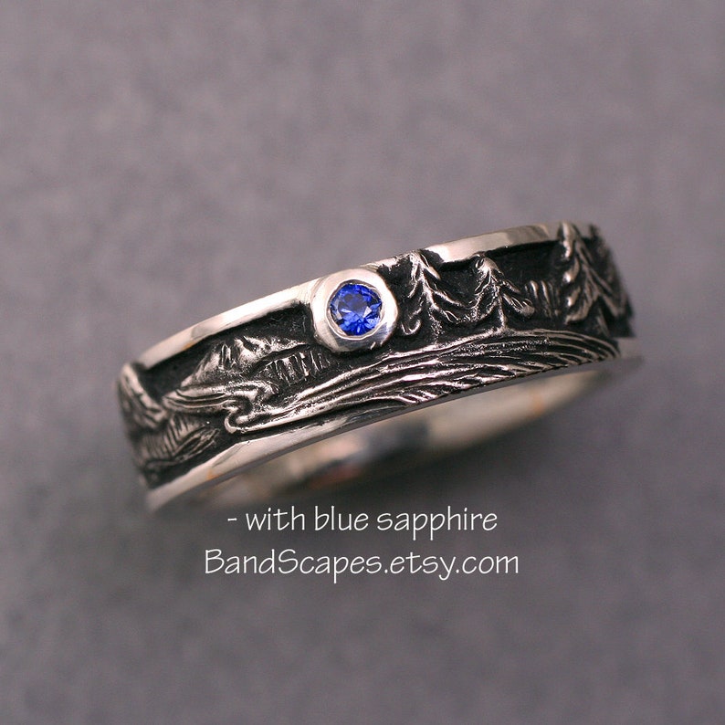 May include: A silver band ring featuring a relief landscape of trees and mountains with a single round blue sapphire set in the metal. The text - with blue sapphire BandScapes.etsy.com is printed below the ring on the gray surface.